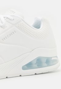 Biały but sportowy z gładką skórzaną cholewką, perforowanym wzorem, widoczną amortyzacją powietrzną w pięcie oraz teksturowanymi detalami.
