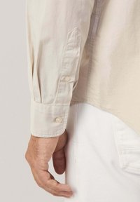 Camicia color beige chiaro con maniche lunghe, polsini abbottonati e orlo leggermente curvato. Tessuto morbido con una leggera lucentezza.