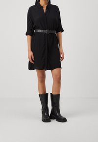 Vestito nero a camicia con bottone frontale, maniche corte arrotolate e vita cinte da una cintura nera. Abbinato a stivali alti neri in pelle.