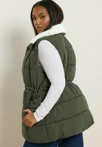 Olive green quilted puffer vest z kremowym kołnierzem sherpa, z regulowanym ściągaczem w talii i bocznymi kieszeniami na zamek, noszony na długim rękawie.