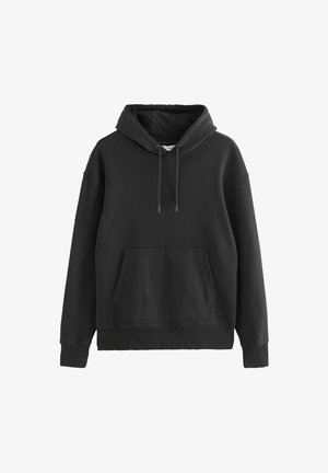 Personne portant un hoodie noir, un sous-vêtement blanc, un jean gris foncé et des baskets noires se tenant devant un fond neutre uni.