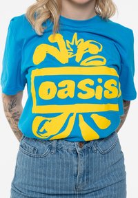 Blue cotton t-shirt featuring bold yellow text "oasis," with abstract designs and short sleeves. Worn with high-waisted denim.