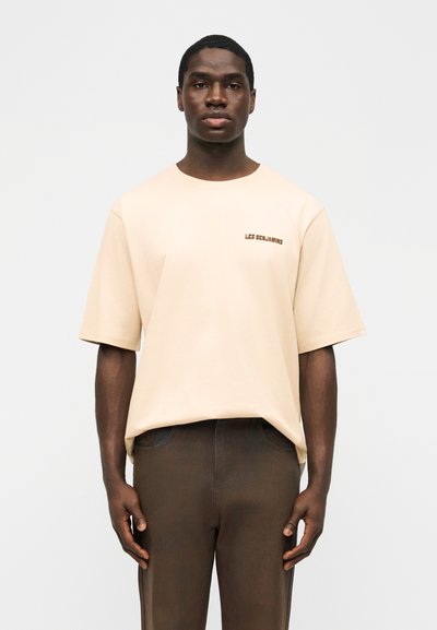 Mężczyzna w beżowej, oversize'owej koszulce z napisem "LES BENJAMINS" i ciemnobrązowych spodniach, stojący na jednolitym jasnym tle.