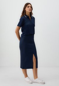Donna in una maglietta blu navy con colletto a maniche corte e una gonna midi abbinata con spacco frontale, che indossa sneaker bianche, su uno sfondo neutro.