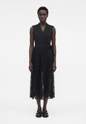 Kvinde iført sort ærmeløs slå-om-kjole med plisseret skørt og blondekant, kombineret med sorte loafers med guldspænder.