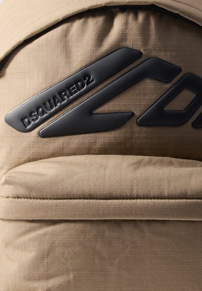 Beige Rucksack aus strukturiertem Stoff, mit einem schwarzen Plastiklogo und markanten, kantigen Designelementen. Reißverschluss oben.