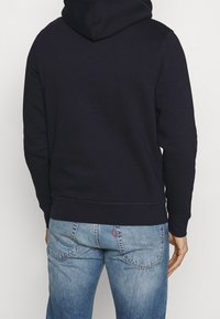 Sudadera de algodón azul marino con capucha y puños acanalados, combinada con jeans de denim claros que tienen una etiqueta roja en el bolsillo trasero.