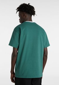 Camiseta de algodón verde de manga corta con cuello redondo y un corte holgado, que presenta aberturas laterales para mayor comodidad y un detalle de costura mínima.