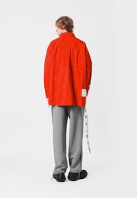 Camisa larga roja con diseño estampado, pantalones de sudadera grises y zapatos negros con una superficie texturada, que muestran un estilo casual.