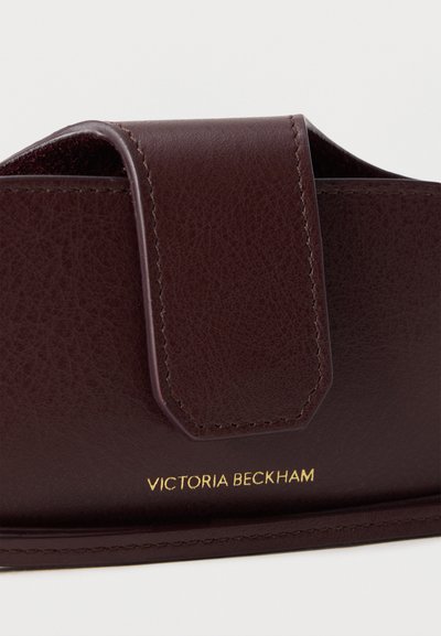 Καφέ πορτοφόλι από δέρμα με υφή, με καπάκι και το όνομα «Victoria Beckham» με χρυσά γράμματα μπροστά σε λευκό φόντο.