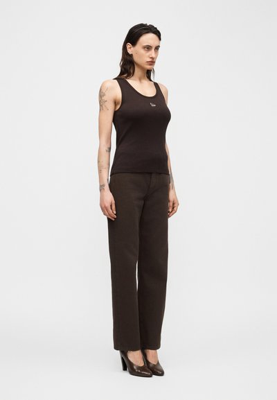 Γυναίκα με μακριά σκουρόχρωμα μαλλιά που φοράει μαύρο αμάνικο τοπ, σκουρόχρωμο παντελόνι με ευθεία γραμμή και καφέ ψηλοτάκουνα παπούτσια, στέκεται μπροστά σε απλό φόντο.