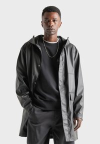 Sort vandtæt jakke med hætte, prydet med trykknapper og en glat tekstur, båret over en sort sweatshirt og matchende bukser.