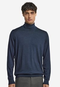 Ung man i en marinblå polotröja och grå byxor, stående mot en enkel vit bakgrund.