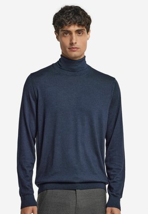 Junger Mann in einem navyblauen Rollkragenpullover und grauen Hosen, der vor einem schlichten weißen Hintergrund steht.