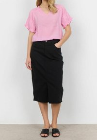 Kvindelig iført en lyserød kortærmet bluse, sort højtaljet midiskirt og sorte åbne slidesandaler, står mod en hvid væg.