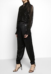 Svart genomskinlig blus med dekorerat mönster, hög krage och långa ärmar. Parat med svarta satinsbyxor med tapered passform och knappdetaljer.