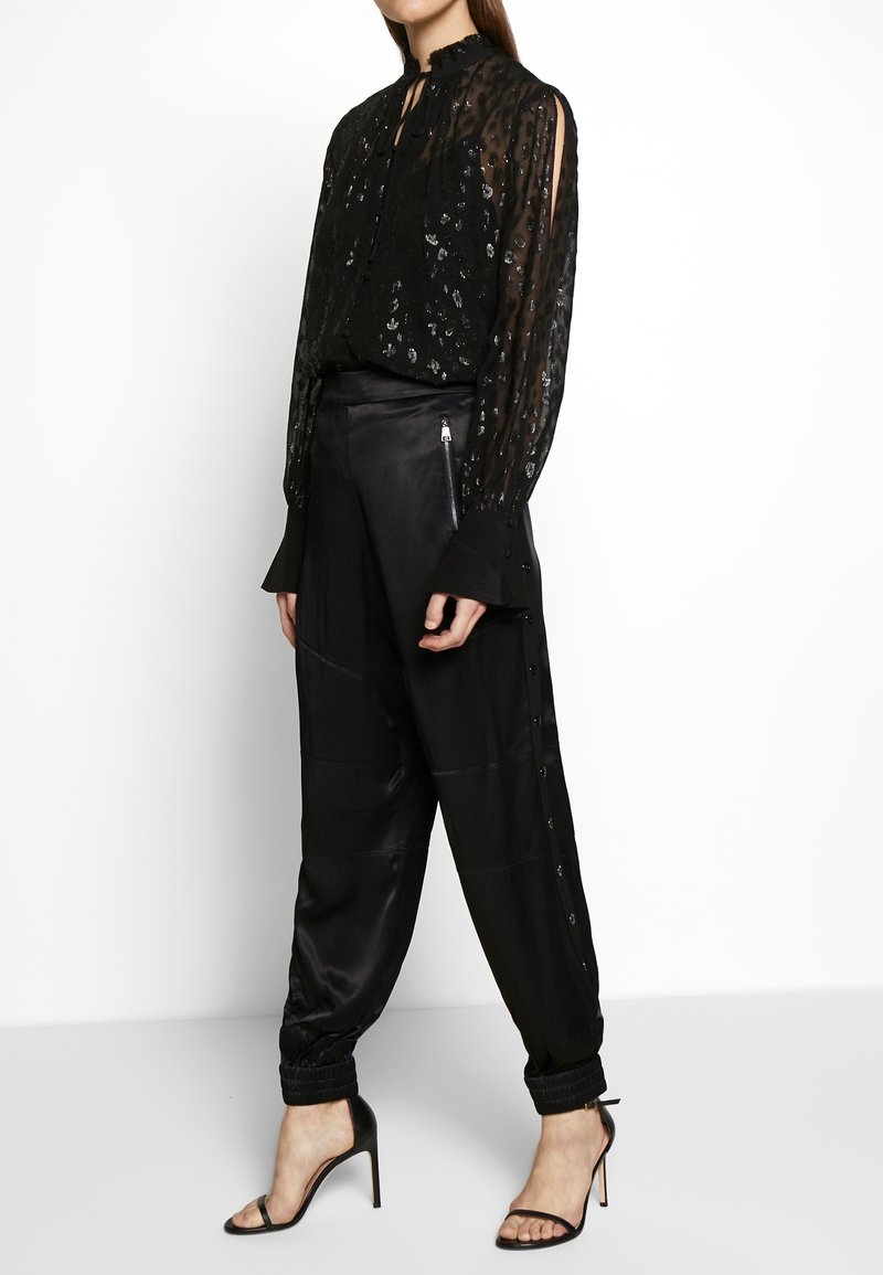 Svart genomskinlig blus med dekorerat mönster, hög krage och långa ärmar. Parat med svarta satinsbyxor med tapered passform och knappdetaljer.