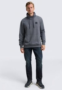 Grauer Kapuzenpullover mit Kordelzug und Fronttasche, aus weichem Stoff. Kombiniert mit dunkelblauer Jeans und grauen Turnschuhen.