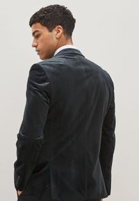 Blazer de terciopelo negro con un ajuste entallado, solapas de punta y puños abotonados. Textura suave, mínima estructura, detalle de ventilación trasera.