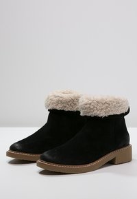 Bottes noires à cheville avec doublure en fausse fourrure beige et semelles en caoutchouc plats marron, placées côte à côte sur une surface blanche.