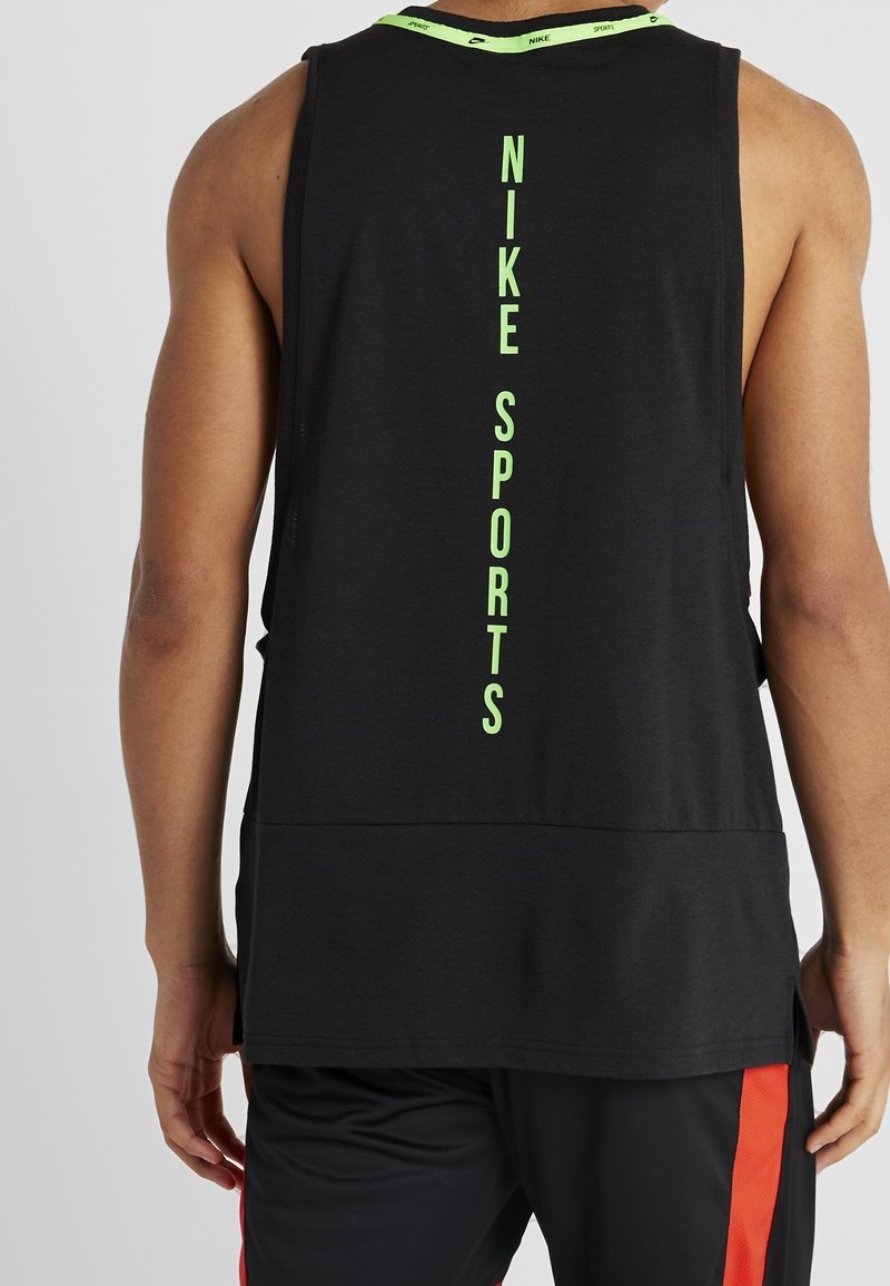 Mies, joka on pukeutunut mustaan ilman hihoja Nike-urheilu toppiin, jossa on vihreä pystysuora logo selässä, ja mustiin shortseihin, joissa on punainen raita, seisoo selkä kameraan päin.