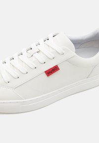 Baskets en cuir blanc avec lacets blancs et une étiquette rouge portant l'inscription "HUGO" sur le côté, sur un fond blanc uni.
