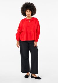 Blusa rossa con scollatura a chiave e fiocco, abbinata a pantaloni neri a gamba larga. La blusa presenta maniche lunghe e vita arricciata.