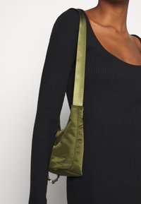 Zielona satynowa torebka na ramię w kształcie trójkąta, o gładkiej fakturze i cienkim pasku. Noszona z czarnym, prążkowanym topem z długim rękawem.