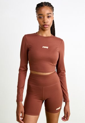 Brown long-sleeve crop top with a round neckline and gold logo, paired with high-waisted matching bike shorts. Smooth, stretchy fabric.