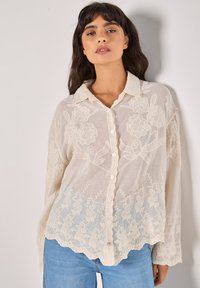 Blusa ricamata color beige chiaro con motivi floreali, chiusura con bottoni frontale, orlo scallop e maniche lunghe, realizzata in un tessuto trasparente e texturizzato.