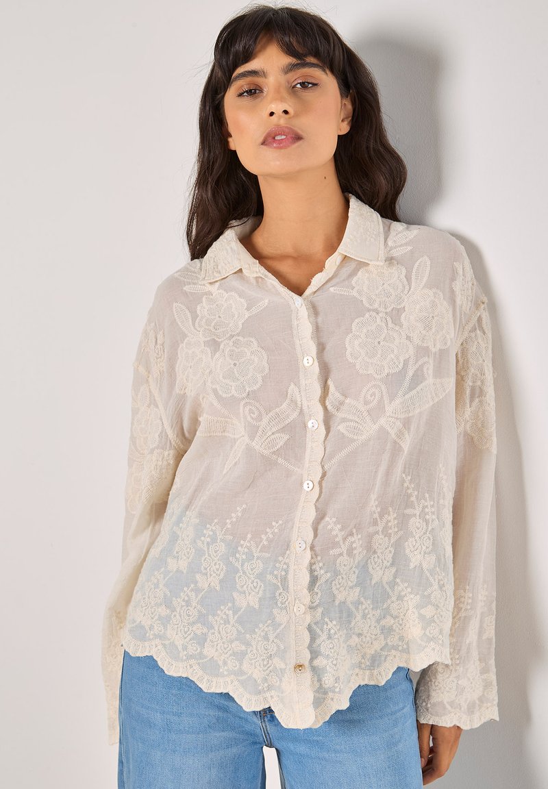 Blusa ricamata color beige chiaro con motivi floreali, chiusura con bottoni frontale, orlo scallop e maniche lunghe, realizzata in un tessuto trasparente e texturizzato.
