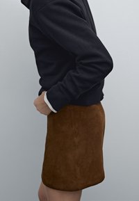 Brun mocka minikjol i kombination med en svart tröja med ribbat detaljer och vita manschetter, visad mot en neutral bakgrund.