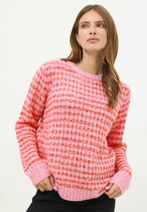 Kobieta w różowo-czerwonym dzianinowym swetrze z prążkowanym dołem i mankietami, pozująca z rękami przy talii na gładkim tle.