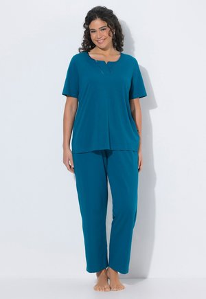 Donna con capelli ricci sorridente, in piedi a piedi nudi con un top a maniche corte e pantaloni coordinati di colore verde acqua su sfondo bianco.