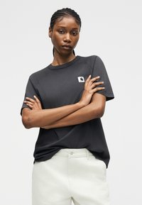 Ung kvinde med fletninger iført sort kortærmet top og hvide bukser, stående med armene over kors mod en ensfarvet baggrund.