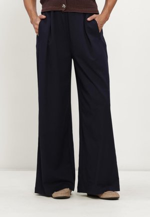 Person wearing loose dark navy wide-leg pants with hands in pockets and beige moccasin-style shoes, standing against a plain background.