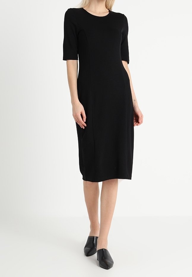 Robe noire longueur genou avec manches courtes, coupe ajustée, tissu lisse et coutures minimales. Portée avec des chaussures noires faciles à enfiler.
