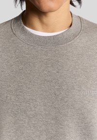 Grå bomullströja med rundad halsringning, med en subtil logotyp längst ner till höger. Texturen är slät och tyget verkar avslappnat.