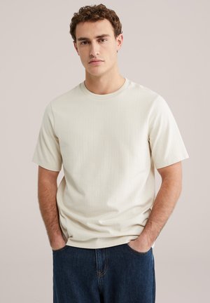 Lichtbeige T-shirt met korte mouwen, ronde hals, geribbelde structuur en een comfortabele pasvorm, gecombineerd met donkerblauwe jeans. Geen zichtbare logo's of ontwerpen.