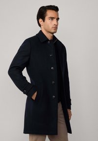 Uomo che indossa un cappotto button-down di lunghezza al ginocchio di un blu navy scuro con colletto, mano sinistra in tasca, rivolto a destra su uno sfondo grigio chiaro uniforme.