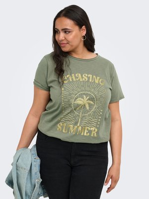 Femeie cu păr lung și închis la culoare, purtând un tricou verde măsliniu cu un desen galben de palmier și textul „Chasing Summer”, ținând o jachetă de denim albastru deschis.