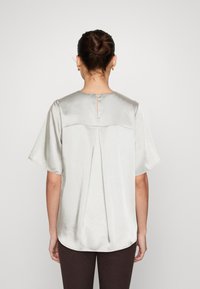 Blouse en satin gris clair avec manches courtes, encolure ronde et ouverture en forme de goutte dans le dos avec un pli pour plus de texture.