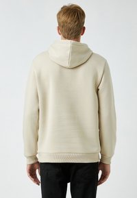 Sudadera beige con una textura suave, capucha ajustable con cordón, puños y dobladillo acanalados. La vista trasera muestra un diseño limpio con un detalle mínimo.