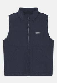 Tmavě modrá vesta bez rukávů s vysokým límcem, předním zipem a dvěma předními kapsami. Má prošívání a malé logo na levé straně.