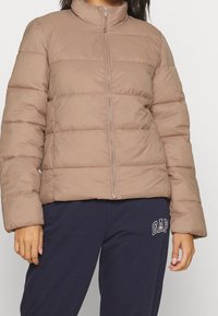 Giacca puffer marrone chiaro con colletto alto, motivo a righe orizzontali e chiusura frontale con zip. Il materiale appare liscio e leggermente lucido.