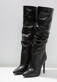 Botas negras de tacón alto hechas de cuero brillante, con una parte superior holgada, punta afilada y detalle de cremallera lateral.