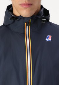 Navy blue K-Way jacket with a high collar, featuring a vertical stripe pattern in yellow, white, and red along the zipper. Logo on chest.