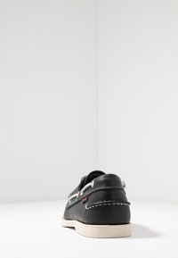 Zapato de cuero negro con costuras blancas, detalles de cordones y suela de color claro. Vista trasera que muestra un diseño curvado y una altura moderada.