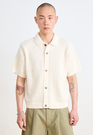 Short-sleeve, knitted cream shirt with a polo collar, vertical eyelet pattern, and textured buttons. Worn with loose-fitting khaki trousers.