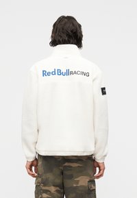 Άτομο που φοράει λευκό φλις μπουφάν με το κείμενο «Red Bull Racing» στην πλάτη και καμουφλαρισμένο παντελόνι cargo, με την πλάτη προς την κάμερα.